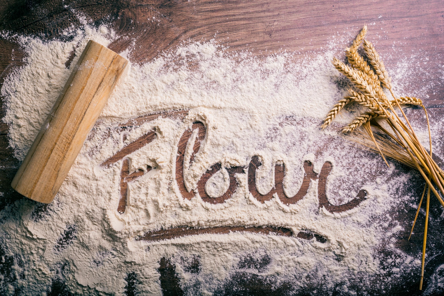 The word 'Flour' written in flour on a dark wooden surface with wheat stalks.